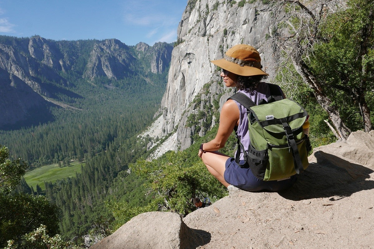 Slacker Day Pack in Olive Green seen on a female hiker on top of a mountain with a valley  running through.